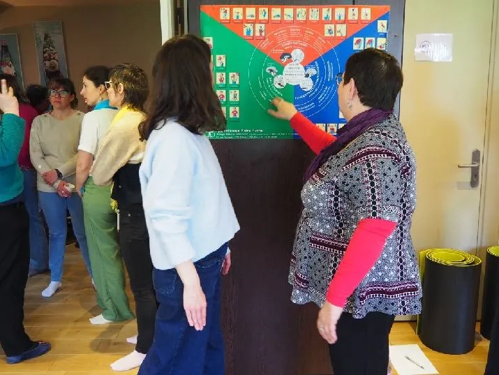 Une formatrice montre une grande planche murale colorée à un groupe de stagiaires debout autour d’elle